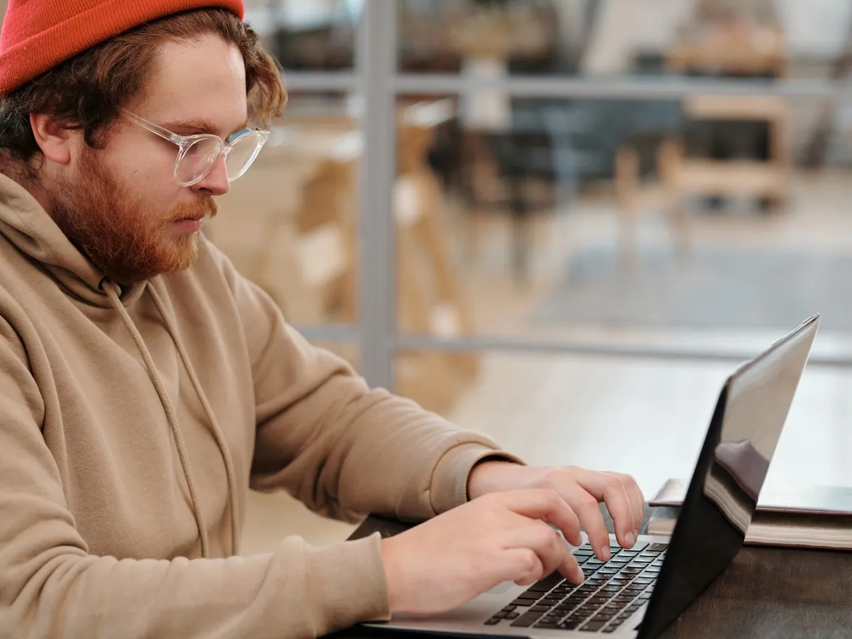 Junger Mann mit Mütze und Brille arbeitet im Café am Laptop